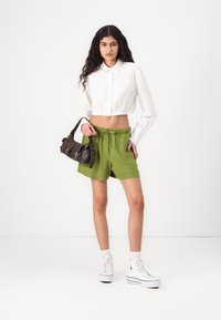 Camisa blanca corta con botones, pantalones cortos verdes con cintura elástica y zapatillas blancas de caña alta. Bolso pequeño oscuro con una correa.