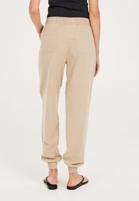 Beige, lockere Anzughose mit elastischem Bund und Bündchen. Mit zwei aufgesetzten Taschen auf der Rückseite und einem weichen, strukturierten Stoff.