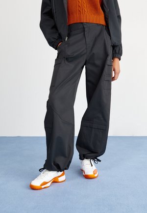 Person iført sorte cargobukser med elastiske manchetter, hvide og orange sneakers samt en orange kabelstrikket sweater under en sort jakke.