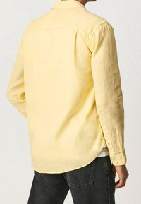 Camisa amarilla de manga larga hecha de tejido ligero, con un collar estándar, cierre de botones en los puños y un dobladillo curvado.