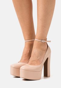 Escarpins à plateforme en cuir verni beige, bout arrondi, bride à la cheville et talon bloc épais et haut. Fini lisse et brillant.