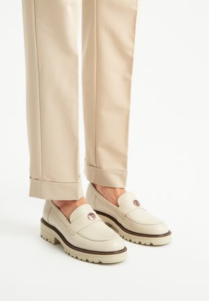 Mocassini in pelle beige con tomaia testurizzata, accentati da un logo rotondo e suola in gomma spessa. Abbinati a pantaloni beige su misura.