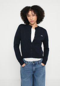 Tumši zils adīts kardigans ar apaļu izgriezumu, pogām priekšpusē, ribotu apakšmalu un aprocēm, ar izšuvumu "2ND" uz kreisā krūškurvja. Kombinēts ar baltu kreklu un zilām džinsa biksēm.