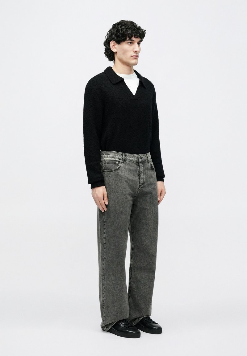 Young man with curly hair stands facing right, wearing black sweater, white undershirt, faded gray jeans, and black sneakers on white background.