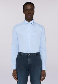 Modrá pruhovaná košile s dlouhým rukávem vyrobená z bavlny, s límcem na knoflíky a malým logem na hrudi, kombinovaná s tmavými džínovými kalhotami.