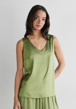 Kvinde med mellemlangt mørkt hår iført en ærmeløs, skinnende olivengrøn V-hals top og matchende løse bukser mod en ensfarvet baggrund.