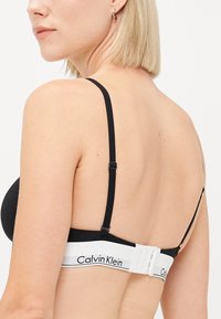 Čierna podprsenka s nastaviteľnými ramienkami a bielym logom Calvin Klein; hladká textúra a priliehajúci strih s uzatváraním na háčik a očko na chrbte.