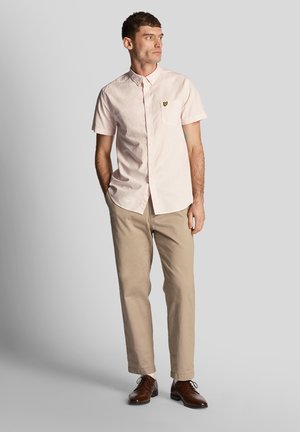Camicia rosa a maniche corte con taschino e logo ricamato, abbinata a pantaloni beige e scarpe eleganti marroni, in piedi su uno sfondo grigio.