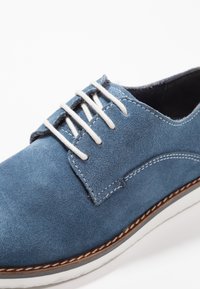 Scarpa di camoscio blu con lacci bianchi, punta arrotondata e cuciture in contrasto. Sole rialzato con un dettagli in beige chiaro. Texture morbida.