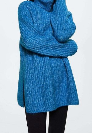 Personne portant un pull en maille côtelée bleu à manches longues avec des fentes latérales, associé à un pantalon noir sur un fond clair.