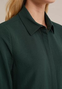 Ciemnozielona koszula z kołnierzykiem o luźnym kroju, wykonana z miękkiego materiału, z zapięciem na guziki i subtelnymi detalami szwów.