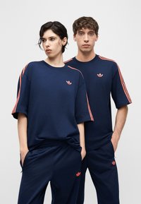 To personer iført matchende marineblå Adidas-treningsdresser med korallfargede striper på ermer og bukser, stående med hendene i lommene.