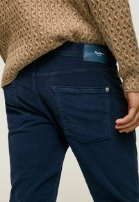 Person som bär marinblå kordflöjtsbyxor med bakfickor och en kamsfärgad kabelstickad tröja, handen vilar vid sidan.