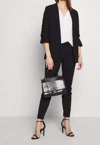 Blazer noir et pantalon avec un haut blanc, tenant un sac à main en cuir crocodile noir. Des escarpins noirs à lanières complètent la tenue.