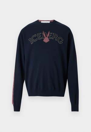 Mann steht und trägt ein marineblaues Sweatshirt mit rotem Design und graue Hose, kombiniert mit schwarzen Schuhen, vor schlichtem weißem Hintergrund.