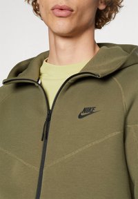 Olive green Nike zip hoodie with a smooth texture, black logo, and contrasting stitching. Features a hood and a full-length zipper.