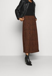 Vrouw in zwarte leren jas, zwarte top, lange rok met bruin patroon en zwarte enkellaarsjes met hakken, staand tegen een effen achtergrond.