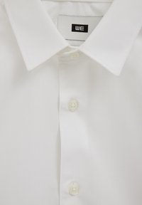 Cămașă albă din bumbac cu guler cu rever, prevăzută cu o fâșie cu cinci nasturi și o etichetă mică neagră cu inscripția „WE” pe interiorul gulerului.