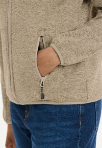 Fleece zip-up jacka i ljusbrun med melerat mönster, med en sidficka och grå detaljer, matchad med blå jeans.