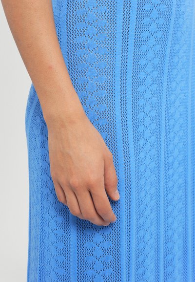 Gaiši zila adīta kleita ar sarežģītām rakstu formām, vertikālām līnijām un mežģīņu līdzīgām dizaina detaļām, izceļot teksturētu apdari un roku darbu.