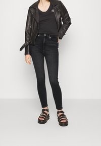 Chaqueta de moto de cuero negro sobre una blusa negra de escote en V, combinada con jeans oscuros de cintura alta y sandalias negras de tiras.