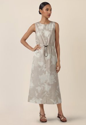 Frau mit zusammengebundenem Haar trägt ein ärmelloses beiges Midikleid mit Blumenmuster, tailliert mit schwarzem Kordelzug, kombiniert mit braunen Sandalen.