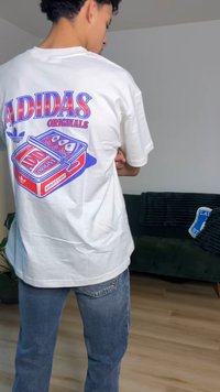 Vit t-shirt med korta ärmar och en färgglad grafisk tryck på baksidan, som visar en retro kassett och Adidas-logotypen.