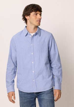 Homme portant une chemise à rayures verticales bleues et blanches avec des boutons et un jean bleu, regardant sur le côté devant un fond uni.