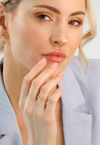 Close-up of a woman with light makeup wearing gold hoop earrings and a gold ring with diamonds, gently touching her lips.