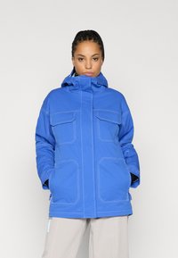 Μπλε αδιάβροχο μπουφάν με χαλαρή εφαρμογή, που διαθέτει υψηλό γιακά, δύο μπροστινές τσέπες, αντίθετο λευκό ράψιμο και ρυθμιζόμενη κουκούλα.