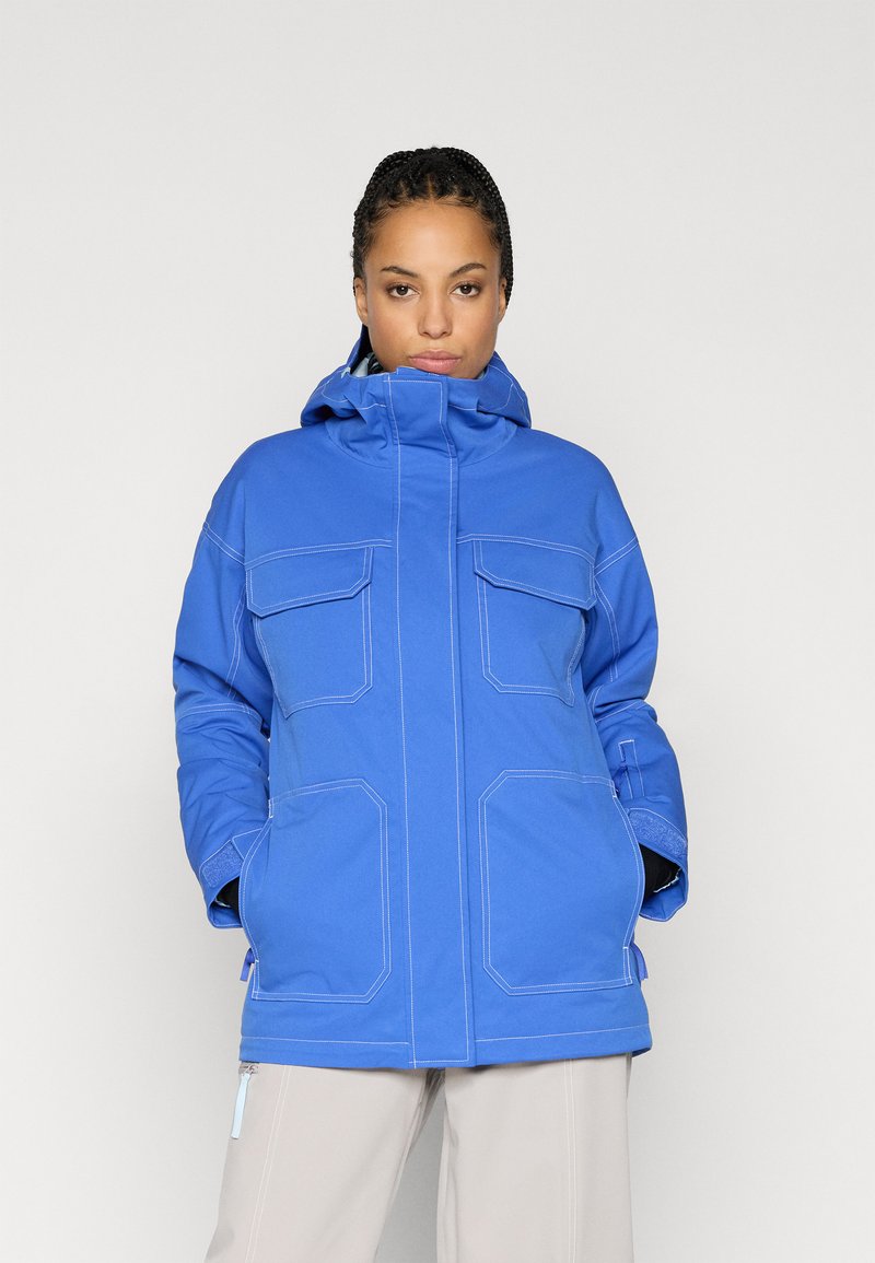 Μπλε αδιάβροχο μπουφάν με χαλαρή εφαρμογή, που διαθέτει υψηλό γιακά, δύο μπροστινές τσέπες, αντίθετο λευκό ράψιμο και ρυθμιζόμενη κουκούλα.