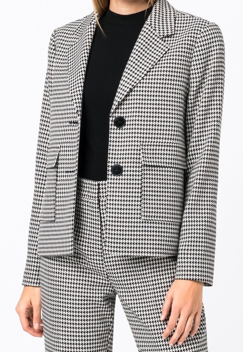Femme portant un blazer à motif pied-de-poule noir et blanc et un pantalon assorti, avec un haut côtelé noir, les mains détendues le long du corps.