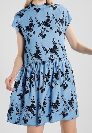 Robe courte bleu clair avec motif floral noir, col montant, manches courtes et jupe froncée portée par une personne aux cheveux blonds.
