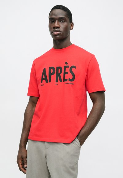 Άνδρας με φωτεινό κόκκινο μπλουζάκι που γράφει "APRÈS" με μαύρα γράμματα και ανοιχτό γκρίζο παντελόνι, στέκεται με το ένα χέρι στην τσέπη μπροστά από απλή φόντο.