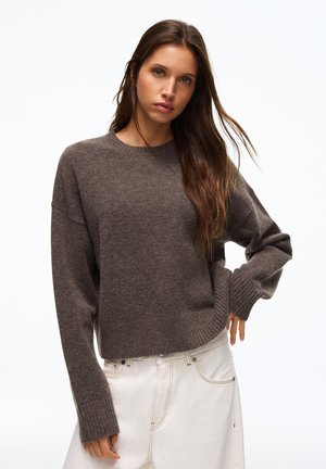 Frau mit langen braunen Haaren, die einen braunen Strickpullover und weiße Hose trägt, posiert mit einer Hand in der Hüfte vor weißem Hintergrund.