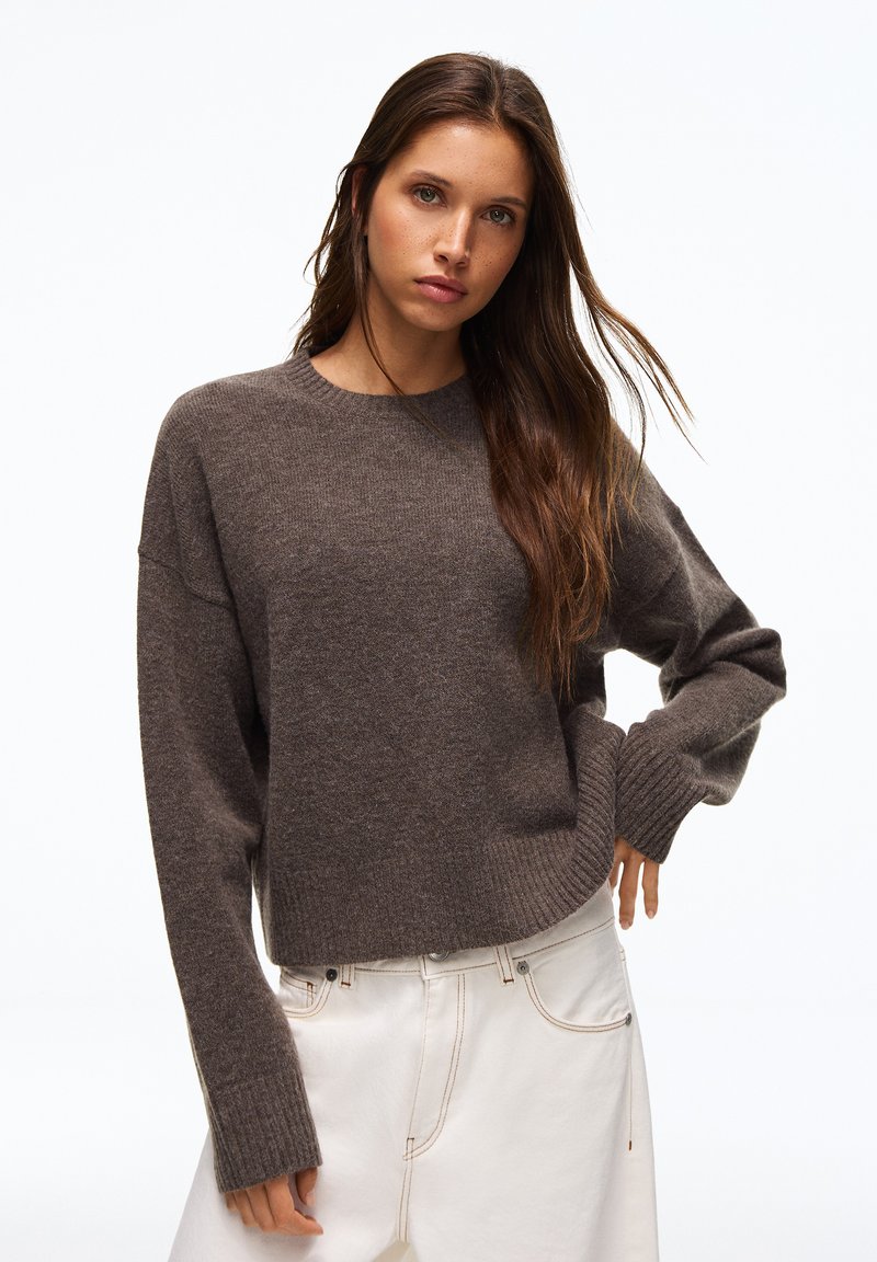 Femme aux longs cheveux bruns portant un pull en maille marron et un pantalon blanc, posant avec une main sur la hanche devant un fond blanc.