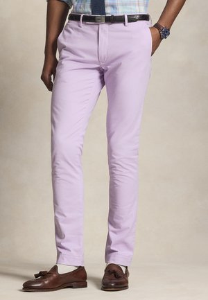 Uomo con pantaloni viola chiaro, mocassini marroni con nappine, calzini bianchi, camicia a quadri, cintura nera e orologio blu, in piedi con una mano in tasca.