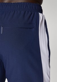 Marineblå atletisk shorts med struktureret stof, prydet med en hvid sidestribe og en lynlås baglomme. Elastisk talje for komfort.