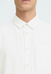 Chemise en lin blanc avec un col à revers, une fermeture boutonnée à l'avant, et une poche poitrine gauche, avec une finition en tissu texturé.