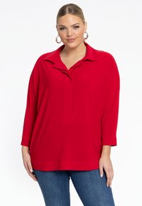 Rote Langarmbluse mit Kragen und teilweisem Knopfverschluss. Weicher Stoff mit lockerem Schnitt, getragen über blauen Jeans. Einfaches Design.