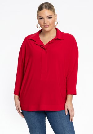 Rode blouse met lange mouwen, een kraag en een gedeeltelijke knopenrij. Zacht materiaal met een ontspannen pasvorm, gedragen over een blauwe spijkerbroek. Eenvoudig ontwerp.