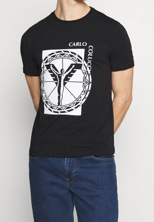 Homme portant un t-shirt noir avec une figure géométrique ailée blanche et le texte "CARLO COLUCCI" sur le devant, associé à un jean bleu.
