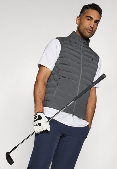 Graue Steppweste mit hohem Kragen, weißes Hemd, dunkelblaue Hose, schwarz-weißer Golfhandschuh, hält einen Golfschläger.
