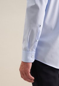 Camicia azzurra chiara con polsini abbottonati, caratterizzata da un ricamo di rosa nera vicino alla manica. Texture liscia e design aderente.