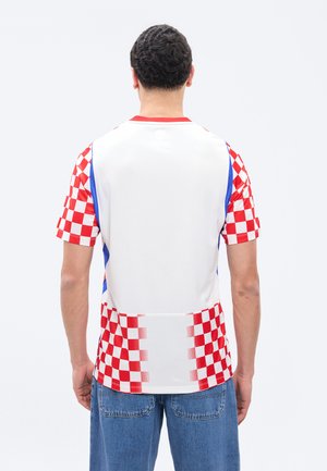 Άνδρας με λευκό πουκάμισο με κόκκινα καρό μανίκια και κάτω πάνελ, στέκεται με την πλάτη προς τα εμπρός, σε συνδυασμό με μπλε τζιν.