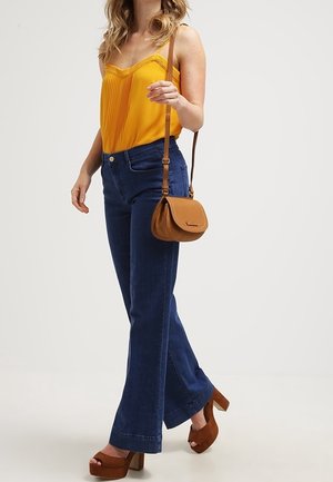 Gele geplooide cami, gecombineerd met donkerblauwe wijde jeans en bruine plateauhakken. Draagt een kleine cognackleurige crossbodytas.