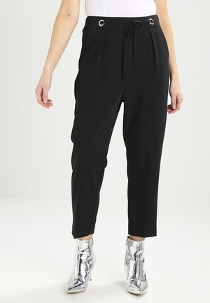 Pantalones negros de cintura alta y corte entallado con pliegues delanteros y cordón, combinados con botines plateados brillantes sobre un fondo claro.