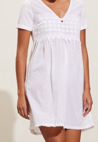 Vit klänning med korta ärmar, prydd med spetsdetaljer och prickig struktur. V-ringning med en knappdekoration och scallop-edged nederkant.