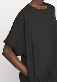 Dark-skinned person wearing a loose black short-sleeve top with their right hand in a side pocket against a plain background.