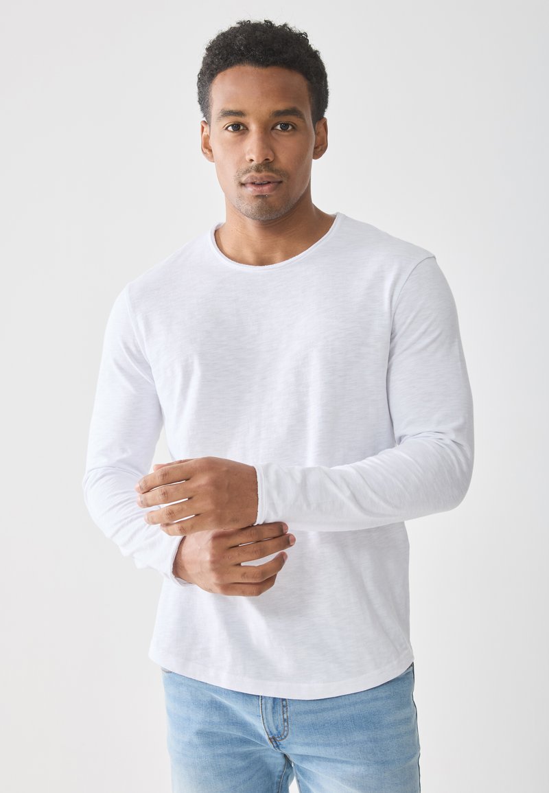 Biała koszulka z długim rękawem z miękkiego, lekkiego materiału o okrągłym dekolcie, zestawiona z jasnoniebieskimi dżinsami. Gładka tekstura.